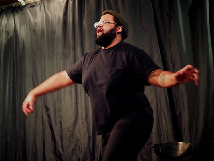 Black man wearing glasses lifts his arms before a black curtain.