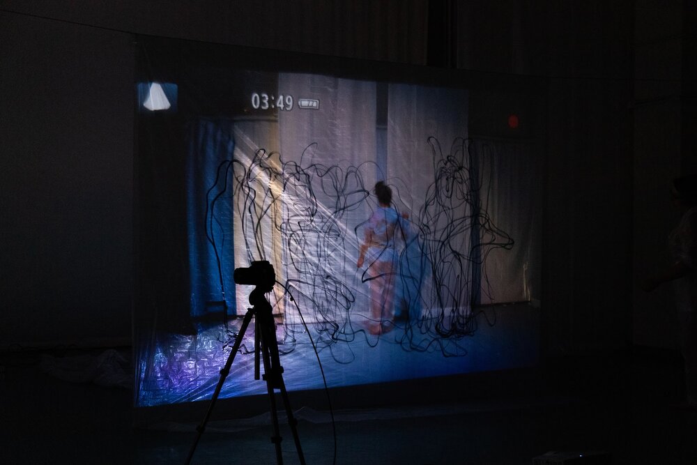 Camera on tripod stands in front of projection with black scribbles and a person in a white jumpsuit facing away