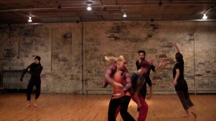 Five people dance in a room with a wooden floor and a brick wall.