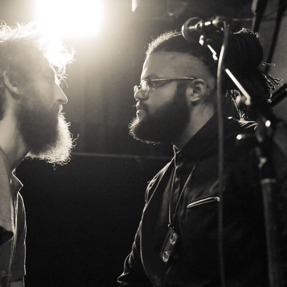 Person with beard and Black man look each other in the eye, in front of microphone on stand.