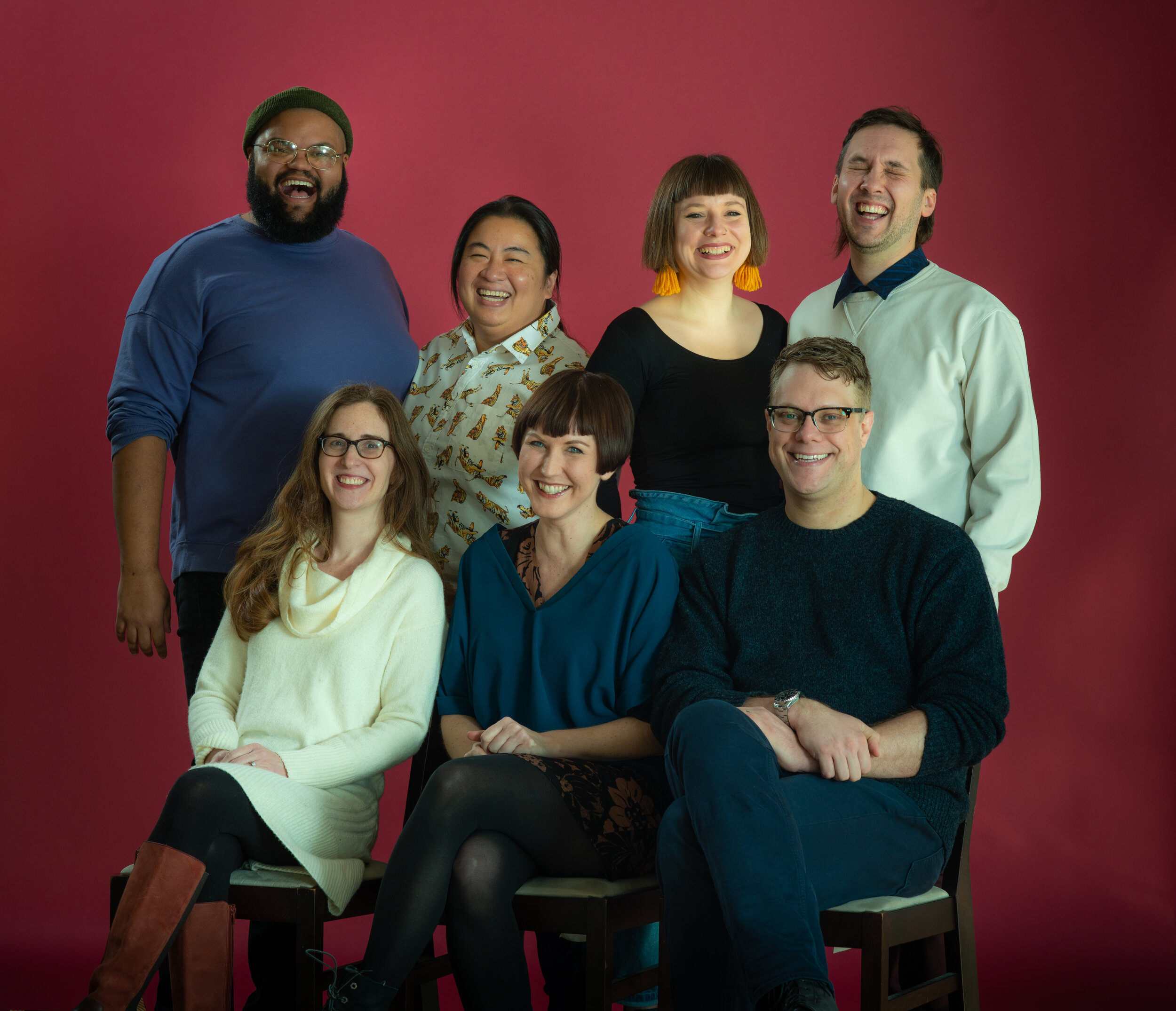 A diverse group of seven people in two close rows face the camera, smiling and laughing.