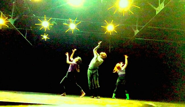 Three people arch their backs away from each other and make claws with their hands, with green and yellow theater lights above them.