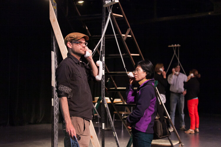 Two people cradle white vintage telephones to their ears, standing before a step ladder. A group of three people hold phones to their ears in the background.