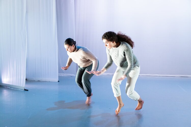 Two people with light skin and brown hair crouch with fingers pressed upwards in white box theater.