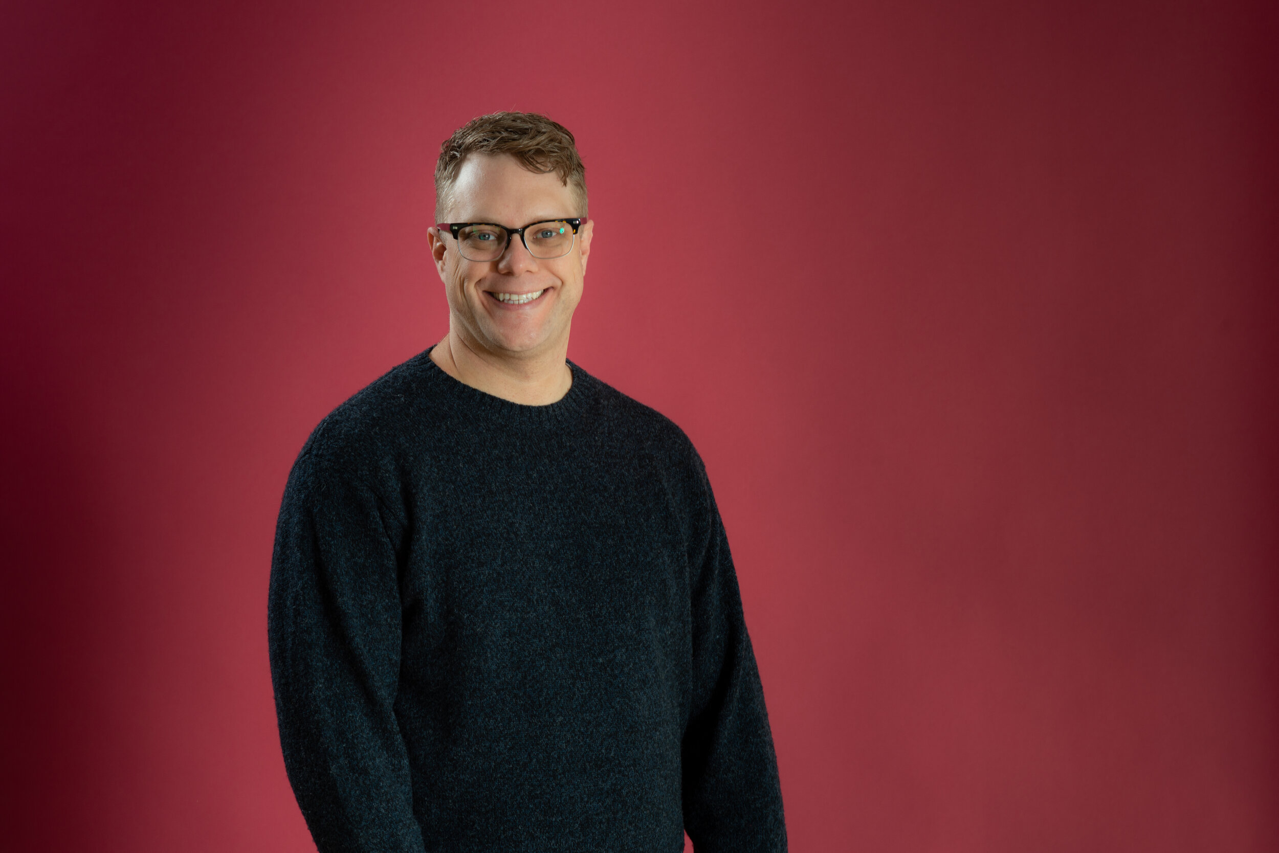 White person with blond hair, blue eyes, and glasses smiles before a red background.