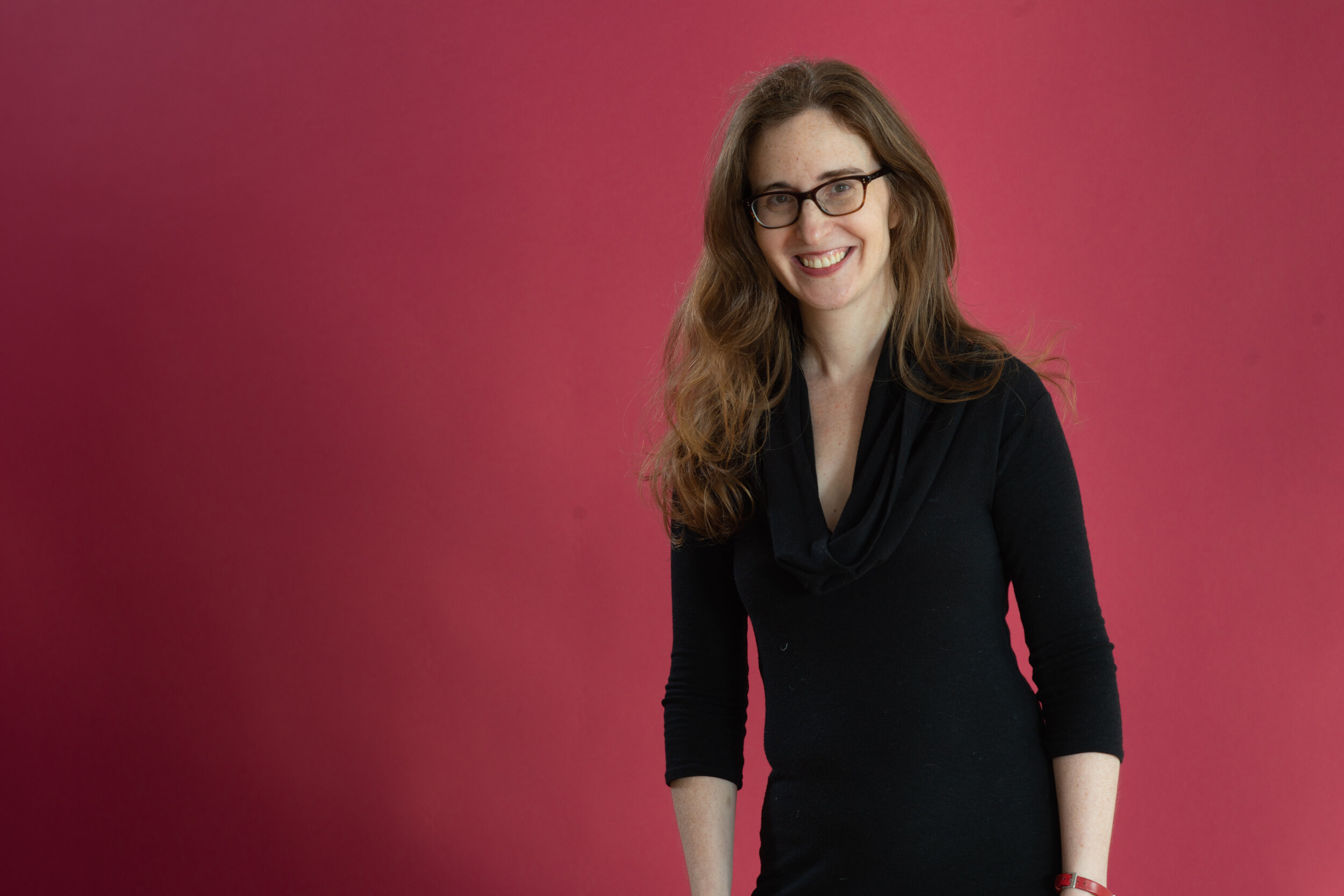 Woman with light skin, red hair, and glasses smiling in front of red background.